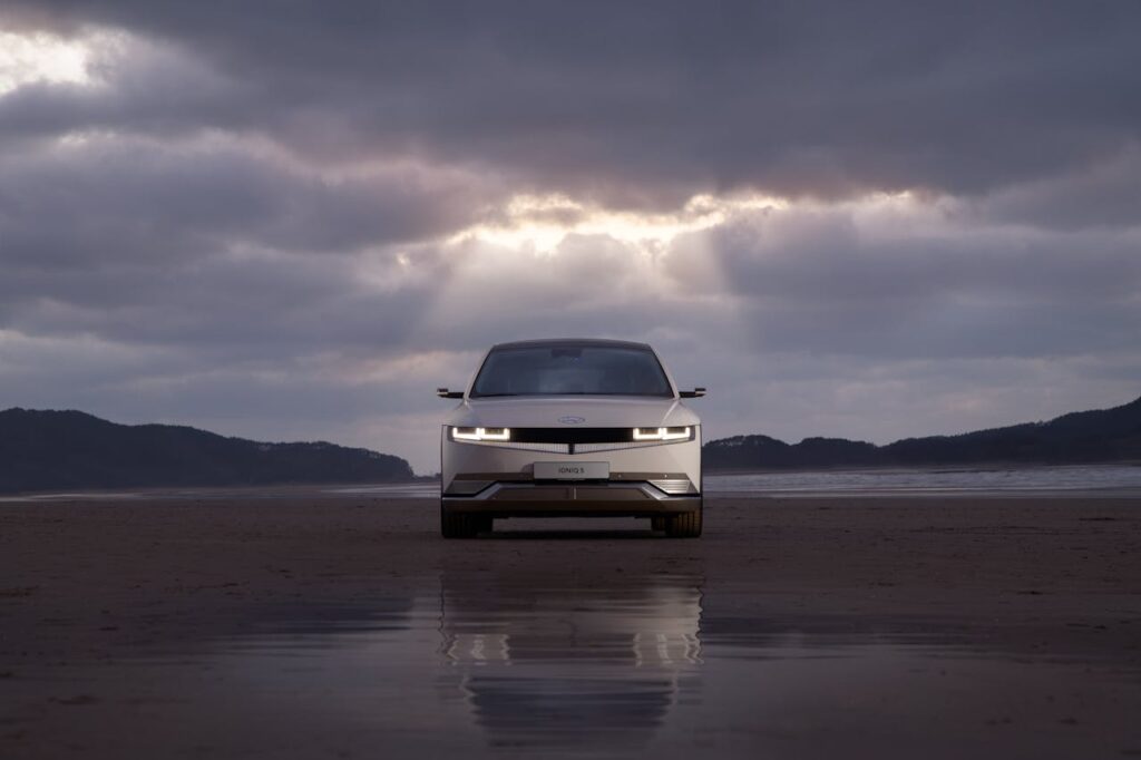 A white electric car reflecting on a wet road under a cloudy sky with sunbeams.